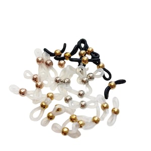 May include: A close-up of a pile of clear plastic eyeglass strap holders with gold, silver, and rose gold coloured metal beads. The strap holders are designed to keep glasses from slipping off the ears.