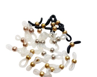 Puede incluir: Primer plano de un montón de soportes de correa para gafas de plástico transparente con cuentas de metal de color dorado, plateado y oro rosa. Los soportes de correa están diseñados para evitar que las gafas se deslicen de las orejas.
