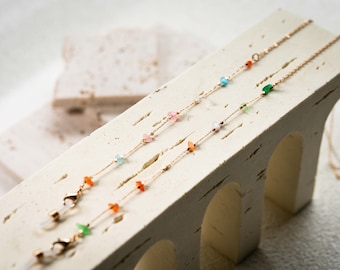 May include: Three gold chains with small colorful gemstone beads. The chains are displayed on a white textured surface.