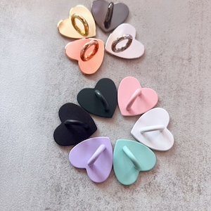 May include: Assortment of heart-shaped phone ring holders in various colors, including black, white, pink, purple, green, gold, silver, and rose gold.