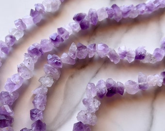 Op de afbeelding: Een streng amethyst edelsteen kralen, elke kraal is een ruwe, natuurlijke kristal met een paarse tint. De kralen zijn aan elkaar geregen op een doorzichtige draad.