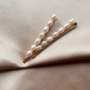 Può includere: Due fermagli per capelli dorati con dettagli di perle bianche. I fermagli sono disposti a forma di V su uno sfondo di tessuto beige.