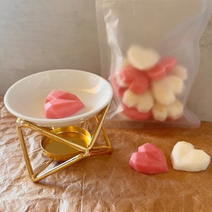 May include: A gold metal wax melt warmer with a white ceramic dish, holding two pink heart-shaped wax melts. A clear bag contains more heart-shaped melts. Two heart-shaped wax melts, one pink and one white, are on the surface.