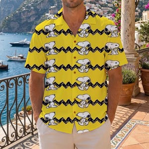 May include: A bright yellow short-sleeved button-down shirt featuring a repeating pattern of the cartoon character Snoopy in a sitting pose, interspersed with horizontal black zigzag lines across the entire fabric of the garment.