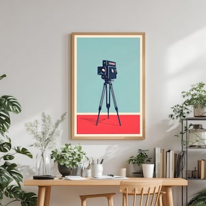 May include: A framed art print depicting a vintage camera on a tripod, set against a teal and red backdrop. The artwork is displayed on a wooden desk with a wooden chair, surrounded by plants. The print has a retro, minimalist style.
