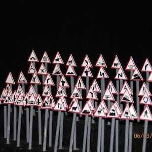 Puede incluir: Una colección de señales de tráfico en miniatura con varios símbolos en blanco y negro sobre fondo blanco con bordes rojos. Las señales están dispuestas en forma triangular sobre una superficie negra.