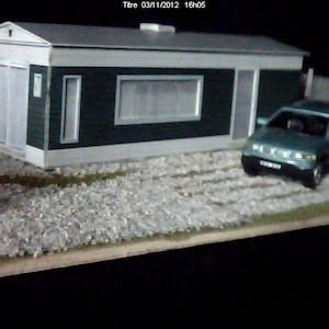 May include: A small green model house with white trim and a gray roof sits on a bed of white gravel. A blue car is parked in front of the house.