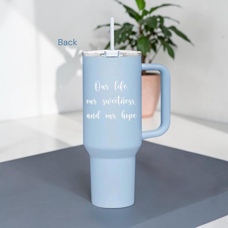 Op de afbeelding: Een lichtblauwe ge&iuml;soleerde beker met een handvat en een rietje. De beker heeft de tekst "Our life, our sweetness, and our hope" in witte letters. Ontworpen om drankjes koud of warm te houden, ongeveer 900 ml.