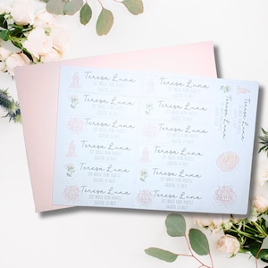 May include: A sheet of address labels with the name "Teresa Luna" printed in a script font. The labels have a light blue background with floral and religious illustrations. A pink envelope is behind the labels.