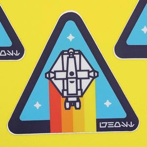 Peut inclure: Un autocollant triangulaire bleu et blanc avec un vaisseau spatial volant à travers un arc-en-ciel. L'autocollant porte le texte "LEANN".