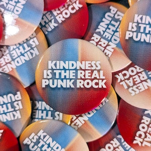 Peut inclure: Plusieurs badges ronds avec l'inscription blanche "KINDNESS IS THE REAL PUNK ROCK". Les boutons affichent un dégradé de couleurs vives, mélangeant le rouge, le bleu et l'orange, pour un effet visuel saisissant.