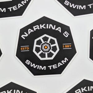 以下が含まれることがあります： NARKINA 5 SWIM TEAMと白い文字で書かれた黒い七角形のステッカーのコレクション。中央にはオレンジ色の点がある白い幾何学的なエンブレムがあり、デザインの両側にオレンジ色の文字でESTDと5 BBYが配置されています。