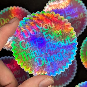 K&ouml;nnte beinhalten: Ein holografischer Aufkleber mit dem Text "Do You Guys Ever Think About Dying?" in einem Regenbogen-Farbschema.