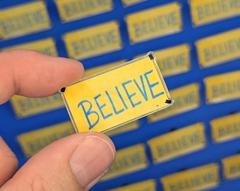 May include: A rectangular acrylic pin featuring a yellow background with the word BELIEVE written in blue capital letters. The pin has small black corner accents and is held against a blue surface covered in identical yellow pins.