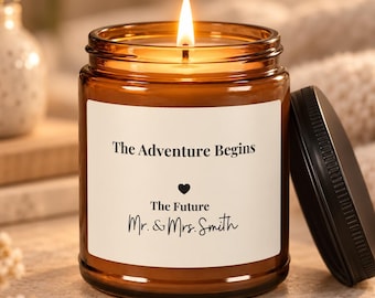 May include: A lit candle in an amber glass jar with a black lid. The white label features the text The Adventure Begins, a small heart icon, and the text The Future Mr. & Mrs. Smith in black ink. The candle flame is bright and centred.