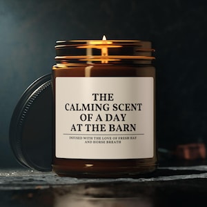 May include: A brown glass jar candle with a lit wick and a white label. The label features black text that reads: THE CALMING SCENT OF A DAY AT THE BARN, INFUSED WITH THE LOVE OF FRESH HAY AND HORSE BREATH. A black metal lid rests beside the candle jar.