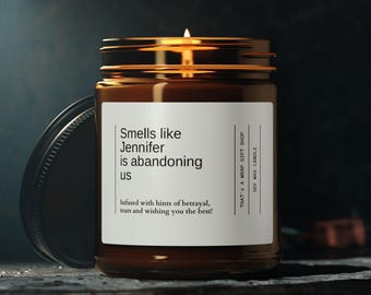 May include: A brown glass candle jar with a white label that reads "Smells like Jennifer is abandoning us. Infused with hints of betrayal, tears and wishing you the best!" The candle is lit and the jar is sitting on a dark surface.