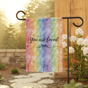 Puede incluir: Una bandera de jardín con un diseño de arcoíris en acuarela. La bandera tiene las palabras "You are loved" en escritura negra. Está colgada de un soporte de metal negro en un jardín.