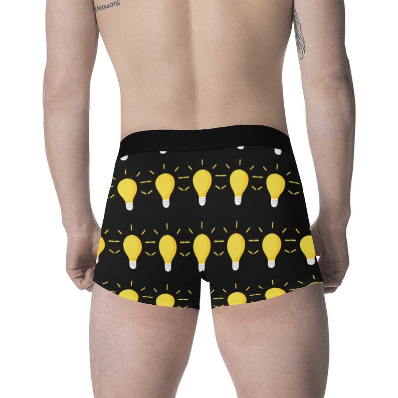May include: Black boxer briefs with a repeating pattern of yellow light bulbs with white bases. The light bulbs have yellow radiating lines. The waistband is black.