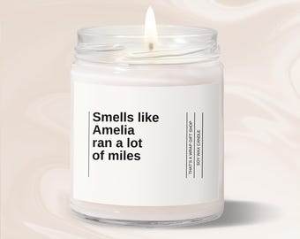 May include: A lit soy wax candle in a clear glass jar. The label reads "Smells like Amelia ran a lot of miles." The text "THAT'S A WRAP GIFT SHOP" and "SOY WAX CANDLE" are also printed on the label.
