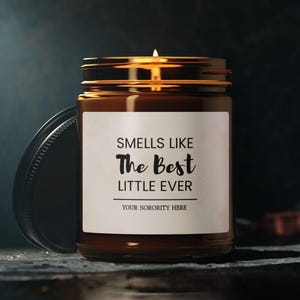 May include: A brown glass candle with a gold lid and a white label that reads "Smells Like The Best Little Ever" and "Your Sorority Here".