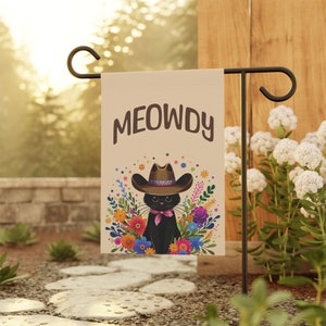 May include: A beige garden flag with a black cat wearing a brown cowboy hat and a red bandana. The cat is surrounded by colorful flowers. The text "MEOWDY" is printed in brown letters at the top of the flag.