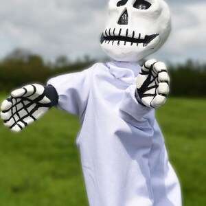 K&ouml;nnte beinhalten: Eine wei&szlig;e Skelettpuppe, die eine wei&szlig;e Robe mit schwarzen Skeletth&auml;nden tr&auml;gt. Die Puppe hat ein bemaltes Sch&auml;delgesicht mit schwarzen Augen und einem schwarzen Mund.