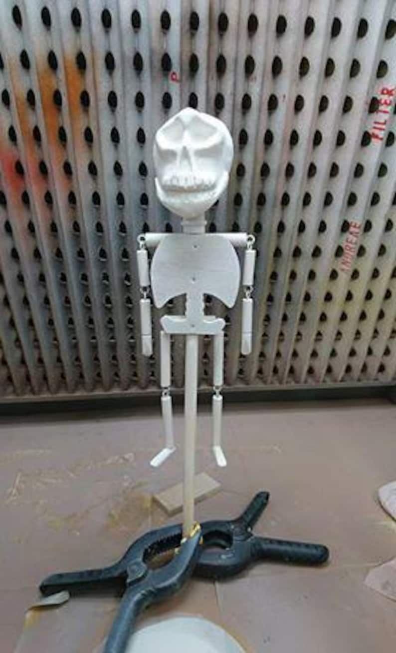 K&ouml;nnte beinhalten: Wei&szlig;e bemalte Holz-Skelettpuppe mit einem l&auml;chelnden Gesicht, befestigt an einem Holzstab und mit einer schwarzen Klemme fixiert.