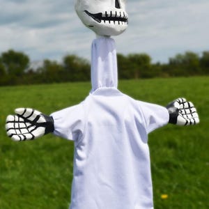 K&ouml;nnte beinhalten: Eine wei&szlig;e Puppe mit einem Sch&auml;delkopf und schwarzen und wei&szlig;en Skeletth&auml;nden. Die Puppe tr&auml;gt eine wei&szlig;e Robe und steht auf einem Feld mit gr&uuml;nem Gras.
