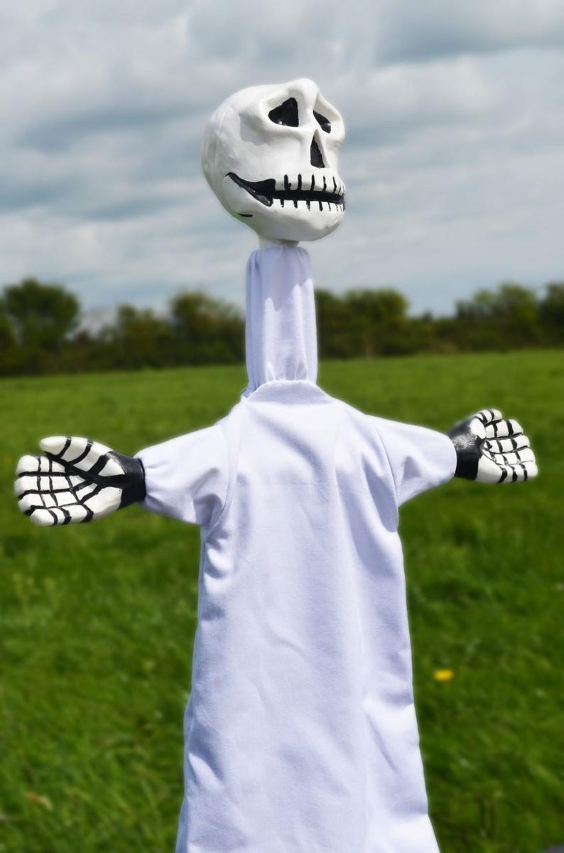 K&ouml;nnte beinhalten: Eine wei&szlig;e Puppe mit einem Sch&auml;delkopf und schwarzen und wei&szlig;en Skeletth&auml;nden. Die Puppe tr&auml;gt eine wei&szlig;e Robe und steht auf einem Feld mit gr&uuml;nem Gras.
