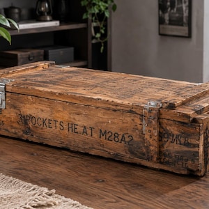 Könnte beinhalten: Eine rechteckige Holzkiste mit verwitterter brauner Oberfläche, ausgestattet mit Metallverschlüssen und seitlichen Seilgriffen. Die Außenseite ist mit schwarzer Schrift versehen: 3 ROCKETS HE AT M28A2, WT 52.5 LB, CU 1.61 und LOADED 5-55.