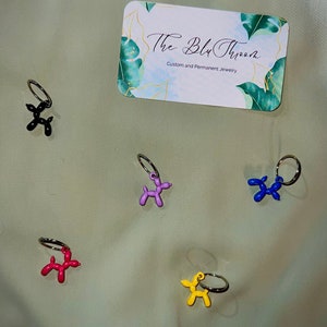 May include: Five balloon animal keychains in different colors: black, purple, blue, red, and yellow. The keychains are shaped like a dog and are attached to silver rings. The keychains are on a light brown fabric background. The text "The Blu Throom Custom and Permanent Jewelry" is visible on a card in the background.