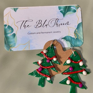 Puede incluir: Un par de pendientes de árbol de Navidad verde con decoraciones rojas y blancas. Los pendientes están hechos a mano y tienen un encanto caprichoso.