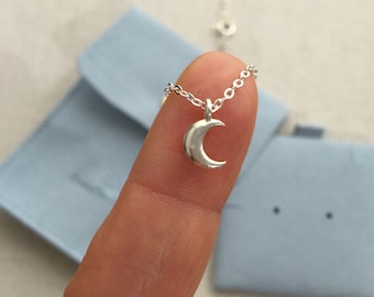 May include: A silver crescent moon pendant necklace. The small, polished moon charm hangs from a delicate chain. The necklace is displayed against a light blue background, suggesting a jewelry presentation.
