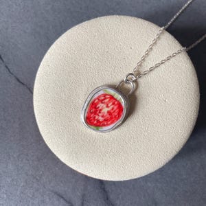 Può includere: Una collana d'argento con un ciondolo a forma di fragola rossa e bianca. Il ciondolo ha un bordo argentato ed è sospeso a una delicata catena d'argento. La collana è esposta su un supporto circolare beige su uno sfondo grigio scuro.