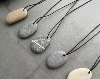 May include: Five necklaces with smooth, oval-shaped stones as pendants. The stones are in shades of gray and beige, and the necklaces are made of brown cord.
