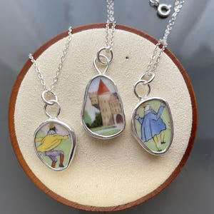 Puede incluir: Tres collares de plata con diferentes colgantes. Los colgantes están hechos de porcelana y presentan un castillo, un hombre con un abrigo azul y un hombre con un abrigo amarillo.