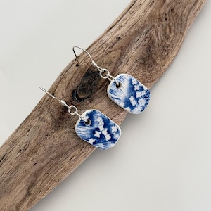 Könnte beinhalten: Ein Paar blau-weiße Porzellanohrringe mit Ohrhaken aus Sterlingsilber. Die Ohrringe haben eine rechteckige Form mit einem blauen Blumenmuster. Die Ohrringe werden auf einem Stück Treibholz vor einem weißen Hintergrund präsentiert.