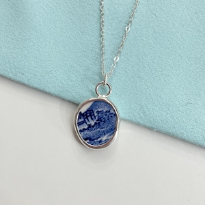 Könnte beinhalten: Eine silberne Halskette mit einem ovalen Anhänger mit blau-weißem Muster. Der Anhänger ist in eine silberne Lünette gefasst, und die Kette ist aus feinem Silber. Das Design auf dem Anhänger scheint eine Landschaft zu sein. Ein klassisches Schmuckstück.