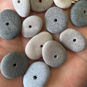May include: A collection of smooth, gray and white stones with holes drilled through the center. The stones are various shapes and sizes, and they are perfect for crafting jewelry or other decorative items.