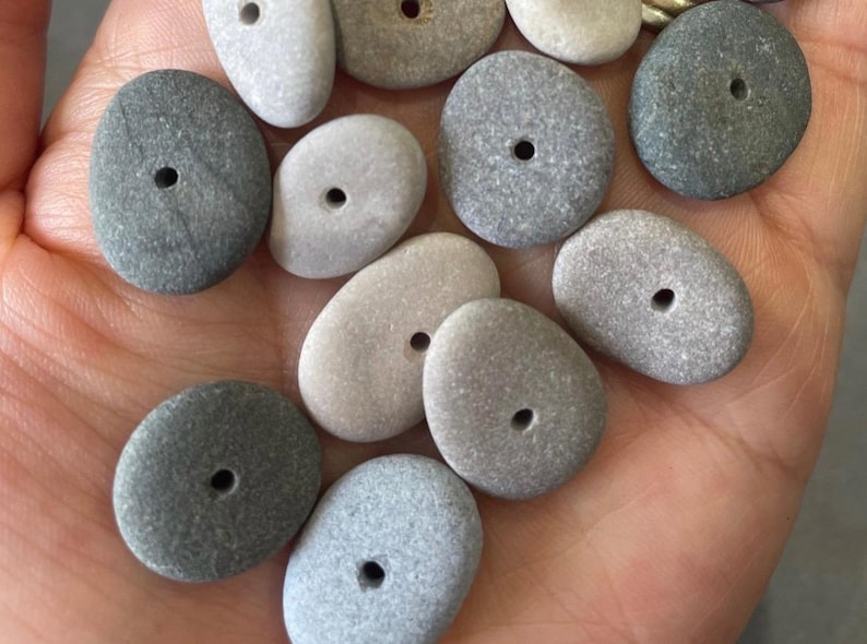 May include: A collection of smooth, gray and white stones with holes drilled through the center. The stones are various shapes and sizes, and they are perfect for crafting jewelry or other decorative items.