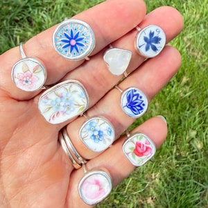 Puede incluir: Una colección de anillos hechos a mano con diseños únicos. Cada anillo presenta un patrón floral o geométrico diferente en tonos de azul, rosa y blanco, engastado en metal plateado. Los anillos se muestran en una mano sobre un fondo de hierba verde.
