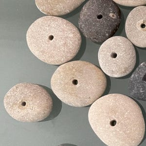 May include: A collection of smooth, round stones with holes drilled through the center. The stones are various shades of gray, brown, and white.