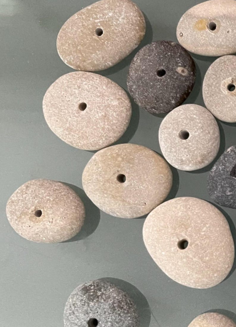 May include: A collection of smooth, round stones with holes drilled through the center. The stones are various shades of gray, brown, and white.