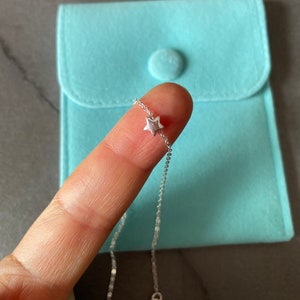 May include: A silver chain necklace with a small star charm. The necklace is on a finger and a light blue velvet pouch is in the background.