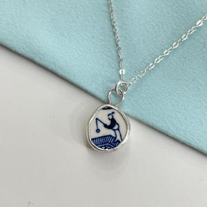 Könnte beinhalten: Eine silberne Halskette mit einem Anhänger, der eine blau-weiße Illustration einer angelnden Person zeigt. Der Anhänger hat einen silbernen Rand und hängt an einer zarten Silberkette. Der Hintergrund ist ein hellblauer Stoff.