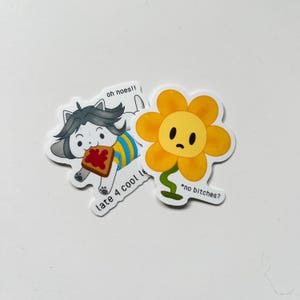 May include: Two cartoon stickers on a white surface. One features a cat-like character with the text "oh noes!!" and "late 4 cool le". The other is a yellow flower with the text "*no bitches?"