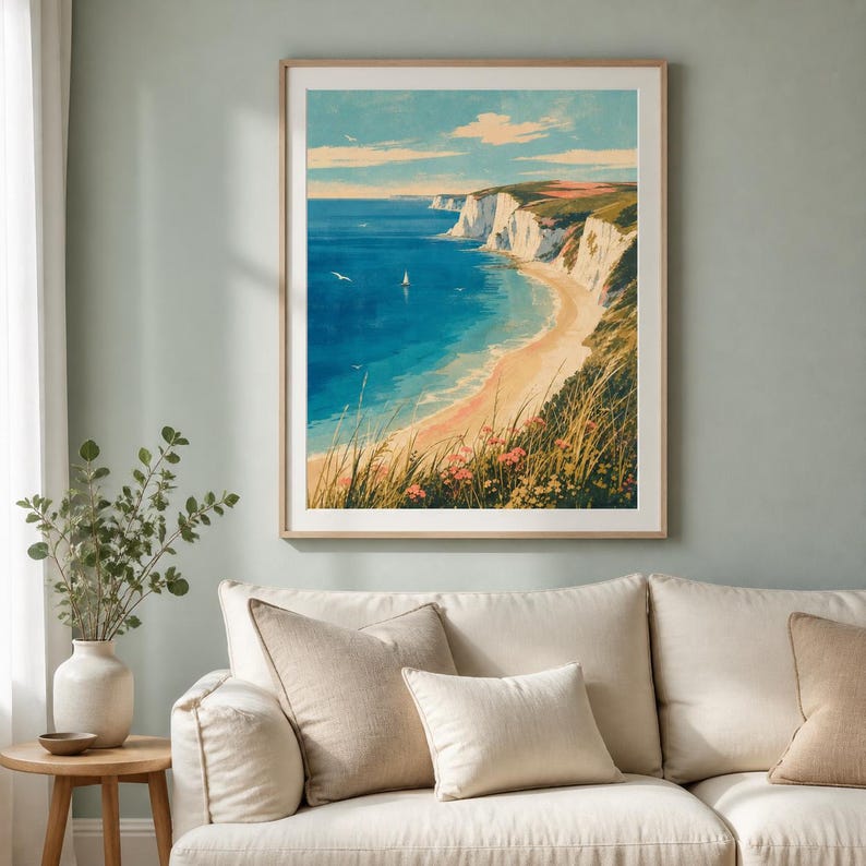May include: A framed wall art print depicting a coastal landscape with white cliffs, a sandy beach, and a deep blue ocean. A small sailboat floats on the water under a light blue sky with soft clouds. The foreground features tall grasses and pink flowers.