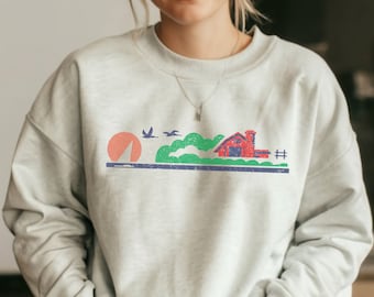 May include: A light gray sweatshirt with a graphic print of a red barn, green hills, and birds flying in the sky. The graphic is in a vintage style with a distressed look.