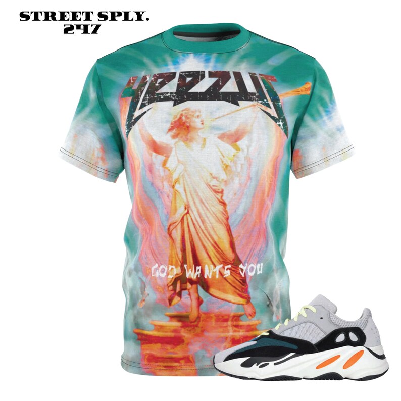 K&ouml;nnte beinhalten: Ein gr&uuml;nes T-Shirt mit einem Batik-Muster, das einen Engel mit Fl&uuml;geln und den Text "God Wants You" zeigt. Das Shirt wird mit einem Paar wei&szlig;er, schwarzer und orangefarbener Sneaker mit einer dicken Sohle kombiniert.